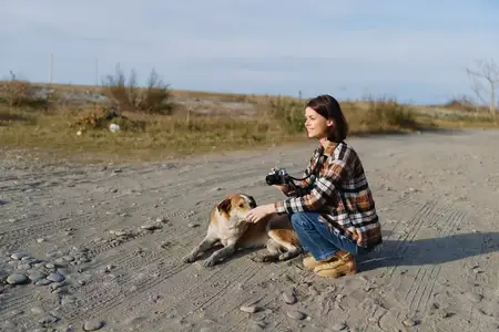 Непринужденная сцена: женщина с камерой разделяет тихий момент с собакой в пустынной пустынной местности, подчеркивая творчество, дружеское общение, исследование природы и спокойное уединение