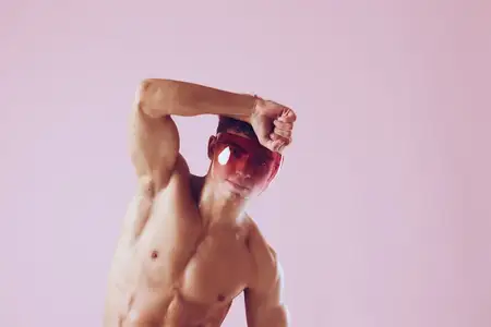 Стильный мужчина-модель с подтянутым телом в солнцезащитных очках принимает игривую позу на фоне мягкого розового фона