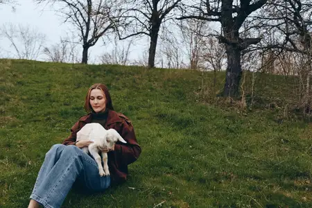 Молодая женщина в коричневой куртке и синих джинсах сидит на зелёной траве, держа белого козлёнка, в то время как голые деревья окружают мирную обстановку на открытом воздухе