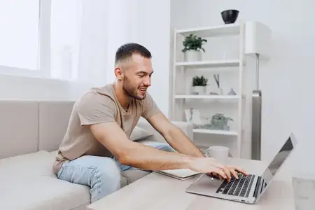 Улыбающийся мужчина сосредоточенно работает за ноутбуком, находясь на удаленке в стильной гостиной