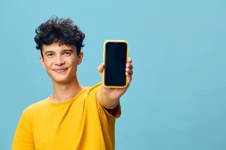 Весёлый молодой человек с кудрявыми волосами держит в одной руке смартфон на фоне мягкого голубого фона