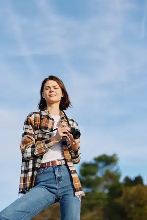 Повседневный портрет молодого фотографа на открытом воздухе: фотоаппарат в руках, джинсы и клетчатая рубашка