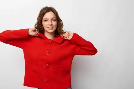 Улыбающаяся молодая женщина в ярко-красном свитере поправляет воротник руками на фоне однотонного белого фона