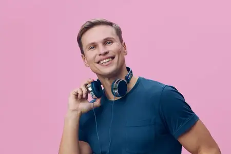 Счастливый мужчина в повседневной футболке и наушниках уверенно позирует с веселым выражением лица на розовом фоне