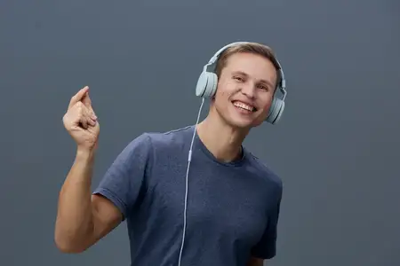 Счастливый молодой человек наслаждается музыкой в больших наушниках, повседневная одежда, дружелюбная улыбка, приподнятое настроение, современный образ жизни, серый студийный фон, позитивная…