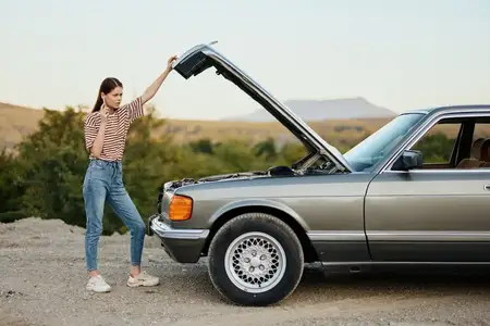 Женщина стоит рядом со сломанным, опасным старым автомобилем с открытым капотом, держа в руке гаечный ключ, во время одиночного путешествия по дороге
