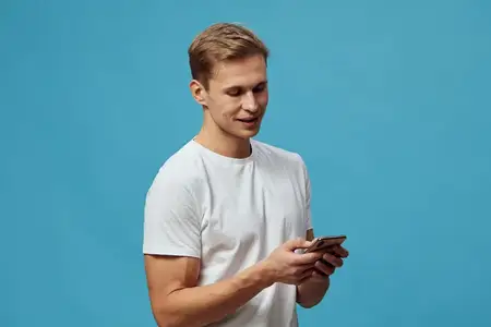 Молодой человек пользуется смартфоном, улыбаясь, в повседневной белой футболке, стоит на синем фоне, держит телефон обеими руками, цифровой кочевник общается и социализируется через приложение для…