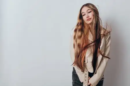 Модель женского пола с длинными каштановыми волосами в бежевой куртке и темных брюках стоит с закрытыми глазами и спокойным лицом на нейтральном фоне для фото в повседневном стиле