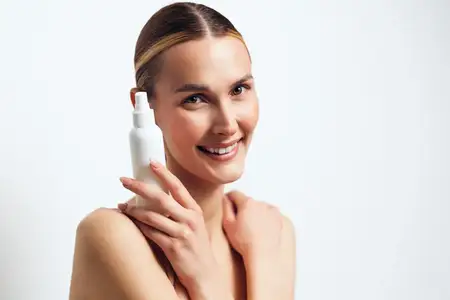 Молодая женщина с blond-волосами мягко улыбается, держа белый косметический флакон-спрей близко к лицу на фоне чистого белого фона, демонстрируя естественную красоту и заботу о себе