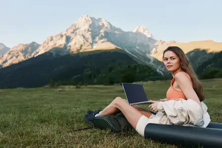 Спокойная сцена: женщина работает за ноутбуком, отдыхая на коврике в огромном альпийском лугу, окруженном скалистыми вершинами