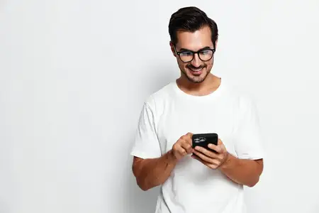 Веселый молодой человек в белой футболке и очках увлечен своим смартфоном
