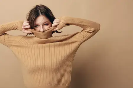 Улыбающаяся женщина в бежевом вязаном свитере закрывает нижнюю часть лица воротником, глаза видны