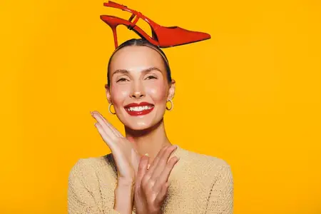 Счастливая женщина с красной туфлей на высоком каблуке на голове улыбается с закрытыми глазами на ярком оранжевом фоне