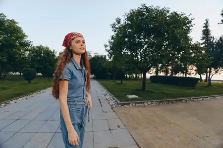 Женщина в стиле современного бохо-вестерн деним с красной банданой уверенно позирует возле зеленых деревьев на мощёной дорожке, выделяясь на фоне теплых тонов цветопередачи плёнки