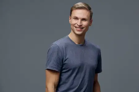 Улыбающийся молодой светлокожий мужчина в голубой футболке смотрит в камеру, изолирован на сером фоне, повседневный стиль, уверенный, счастливый, позитивный, концепция цифрового кочевника, образ…