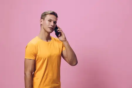 Молодой человек в желтой футболке прижимает смартфон к уху, разговаривает с серьезным сосредоточенным выражением лица, стоит на фоне однотонного розового фона
