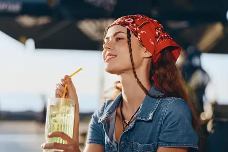 Молодая женщина с красной банданой и в джинсовой рубашке casual пьёт свежий лимонад на улице под солнечным небом, излучая спокойное и освежающее летнее настроение