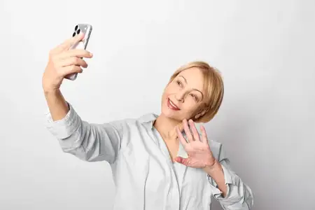 Дружелюбная женщина-блондинка держит смартфон для селфи на ярком белом студийном фоне, улыбается с игривым жестом руки, молодежный образ жизни, общение для социальных сетей