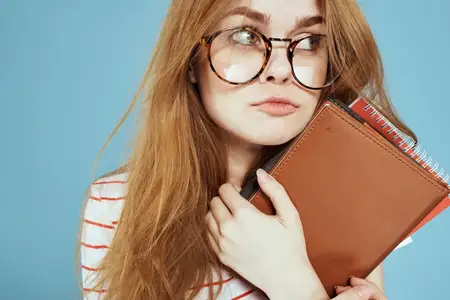 Энергичная студентка с блокнотами и в очках позирует на синем фоне