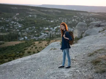 Туристка с рюкзаком за спиной находится в горах на фоне заката и деревьев