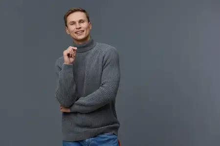 Улыбающийся молодой светлокожий мужчина, одетый в серый вязаный свитер с высоким воротом и синие джинсы, стоит изолированно на сером фоне, держит руку возле лица, счастливое позитивное выражение…