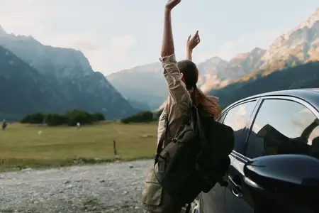 Она одета в зеленую куртку, рюкзак и джинсы, смотрит на далекие вершины, легкий ветерок шевелит ее волосы, создавая естественную, полную приключений атмосферу на открытом лугу, рядом припаркована…