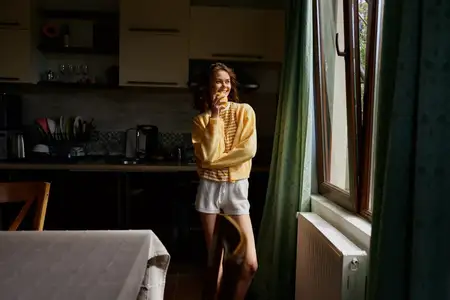 Жизненная сцена с женщиной в теплой кухне у окна, повседневная атмосфера, мягкий дневной свет, расслабленная поза, яркие цвета, повседневная домашняя рутина, комфортное неформальное настроение