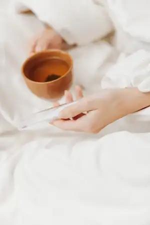 Женщина в постели с чашкой чая и телефоном рядом с миской зелёного чая и ложкой