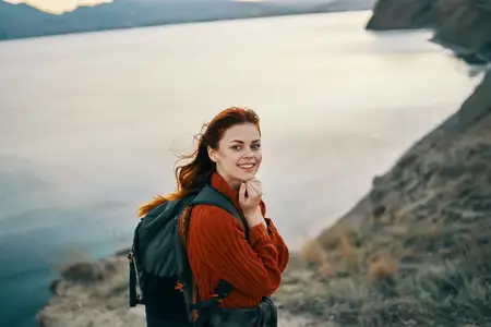 Турист с рюкзаком в горах у моря, в красном свитере, с распущенными волосами, на фоне скал на закате