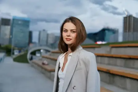 Уверенная бизнес-леди в светло-сером блейзере позирует на открытом воздухе на фоне городского пейзажа в современном профессиональном стиле, при мягком естественном освещении