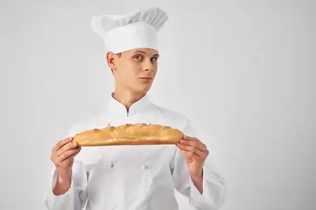 Весёлый пекарь-мужчина откусывает батон, занимаясь приготовлением еды в пекарне