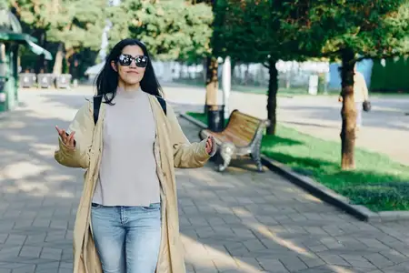 Женщина в солнцезащитных очках, легком пальто и джинсах идет по парковой аллее