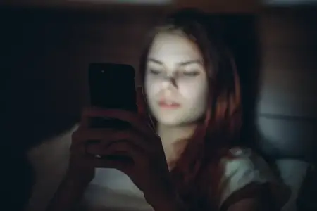 Женщина лежит в комнате на кровати с телефоном в руках, демонстрируя зависимость