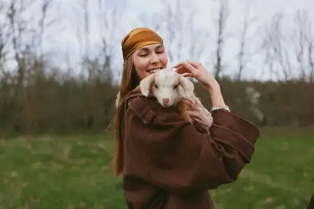 Улыбающаяся женщина в коричневой куртке держит на руках молодого козла на травянистом поле, проявляя нежность и привязанность на природе, подчёркивая здоровую деревенскую жизнь и связь с животными