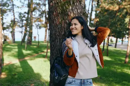 Улыбающаяся женщина на улице у большого дерева в зеленом парке, повседневная одежда и мирная сцена подчеркивают досуг, природное окружение и повседневное оздоровление на свежем воздухе