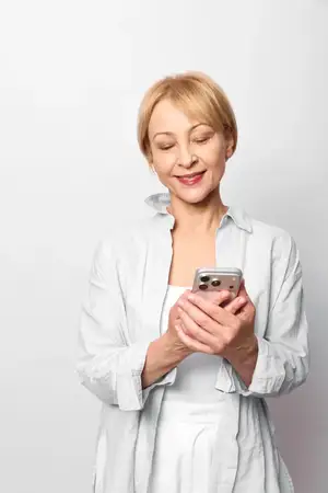 Пожилая женщина с короткими волосами держит смартфон, улыбается в студийном портрете на белом фоне