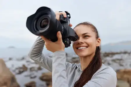 Восторженная молодая женщина с длинными волосами, держащая фотоаппарат и улыбающаяся, снимает живописные виды в прибрежной зоне, отражая радость и творчество в фотографии