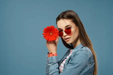 На фотографии высокого качества изображена брюнетка в солнцезащитных очках с красным цветком на фоне голубого фона, передающая атмосферу роскоши и романтики