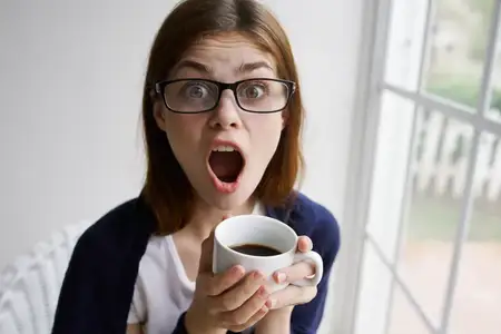 Эмоциональная женщина с открытым ртом держит чашку кофе в интерьере дома у окна во время отпуска