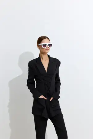 Модная женщина в черном tailored костюме и белых солнцезащитных очках уверенно позирует на минималистичном белом фоне, демонстрируя современный стиль и крутой настрой