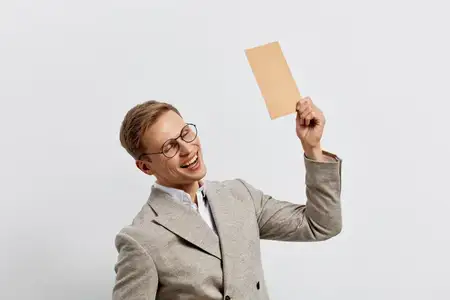 Счастливый улыбающийся молодой светлокожий мужчина в полосатом костюме держит в руке пустую бумажную карточку, изолированный на светло-сером фоне