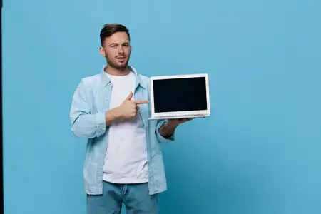 Улыбающийся счастливый загорелый красивый мужчина в повседневной базовой футболке указывает пальцем на новый купленный ноутбук, позируя изолированно на синем студийном фоне