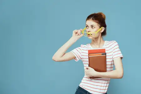 Студентка с книгами в руках на голубом фоне, в жёлтых очках, с модельной причёской, кадрированный вид