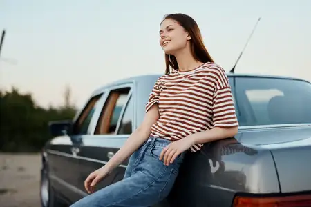 Молодая женщина в полосатой футболке и джинсах отдыхает после вождения, облокотившись на свою машину у дороги, и улыбается, глядя на природу и закат