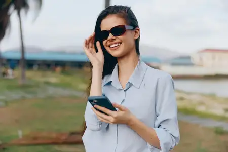 Улыбающаяся женщина в солнцезащитных очках держит мобильный телефон, позируя на фоне живописного водного пейзажа