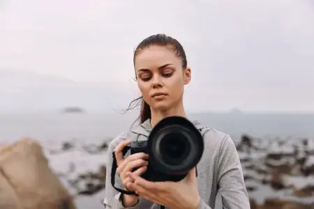 Молодая женщина в серой спортивной одежде сосредоточенно фотографирует океан, что подчеркивает эмоции концентрации и страсти к фотографии на фоне облачного неба