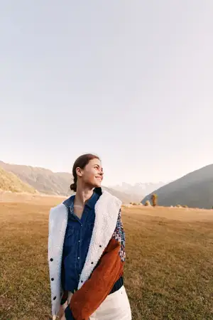 Женщина улыбается на лугу в горах, на ней куртка, осенний портрет на открытом воздухе