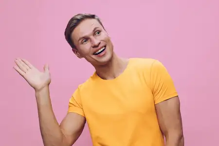 Веселый улыбающийся мужчина машет рукой, одетый в желтую футболку, стоит на розовом фоне с дружелюбным выражением лица, иллюстрируя образ жизни цифрового кочевника, повседневный стиль и позитивное…