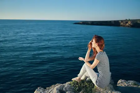 Женщина в футболке и брюках сидит на скале на пляже у моря, вид сверху