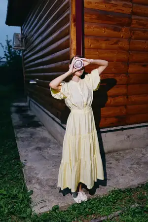 Уличная сцена: женщина-фотограф в желтом платье у деревянного здания в сумерках позирует с камерой, готовая сделать креативный снимок в деревенской обстановке